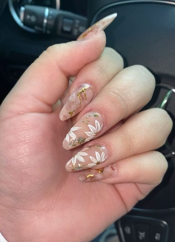 Uñas acrílicas transparentes en forma de almendra con flores blancas y lámina dorada.