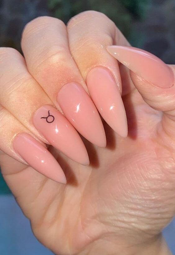 Color de uñas nude con arte minimalista del signo de Tauro en uñas acrílicas largas con forma de almendra