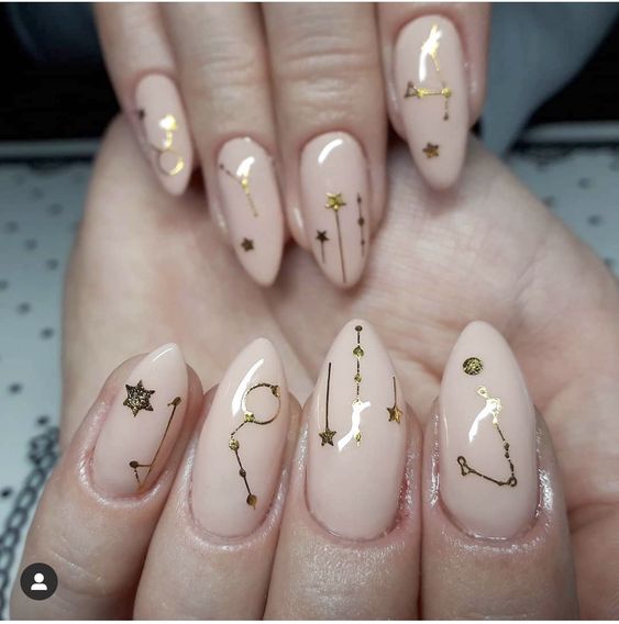 Color de uñas nude con el signo dorado de Tauro y decoración de uñas con elementos celestiales en uñas largas color almendra