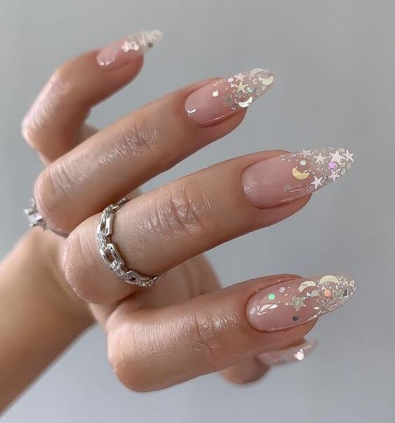 Uñas transparentes en forma de almendra con brillos en forma de estrella y luna en las puntas.