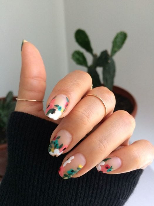 Mano de mujer con uñas con flores coloridas, verde, rosa y blanco mate, sencillas pintadas sobre base transparente para primavera, anillos dorados