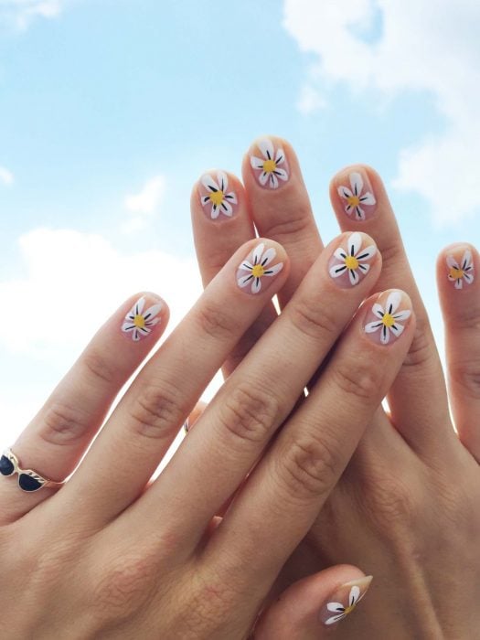 Mano de mujer con uñas con simples flores blancas pintadas que puedes hacer en tu casa para recibir la primavera.  anillo de lentes